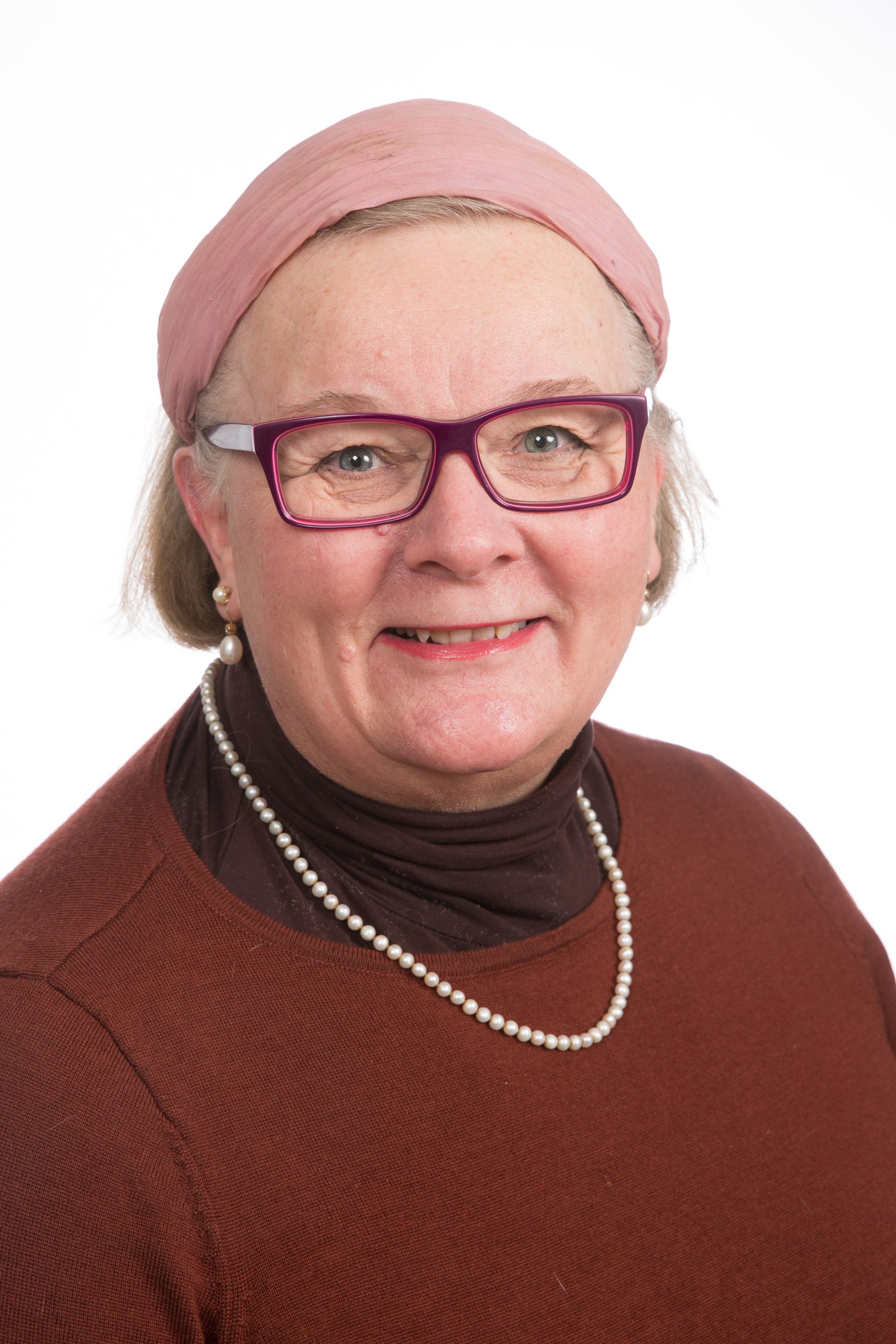 Kuvassa on nainen, jolla on vaalea iho, punasankaiset silmälasit, punainen paita ja kaulassaan helmet sekä hiuksissaan vaalenpunainen panta.